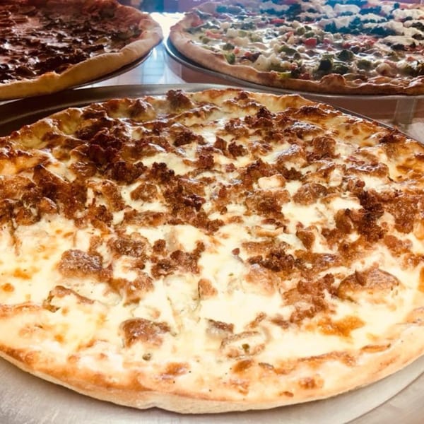 A white pizza with crispy brown sausage, with two other pizzas blurred in the background.