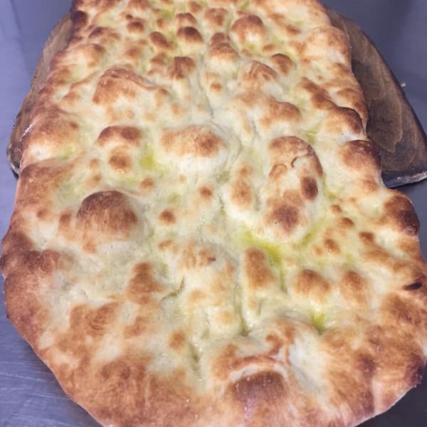 A close-up of a golden-brown focaccia bread, glistening with olive oil, on a wooden peel.