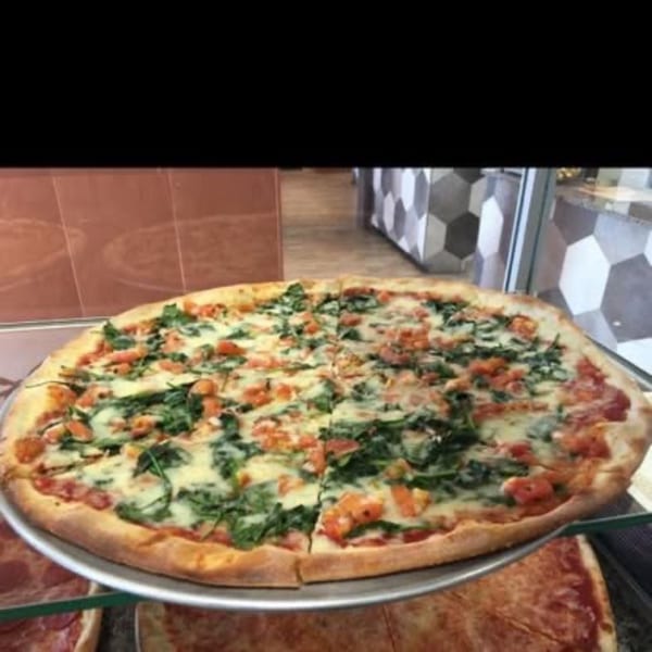 A large, round Pizza Village IV pizza with spinach and diced tomatoes on a metal tray.